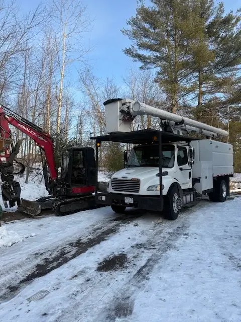 Bucket truck and mini excavator on a snowy job site surrounded by woods