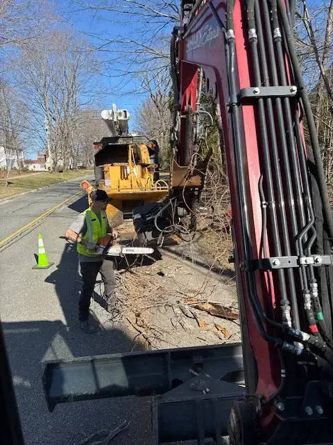 Crew member in a safety vest running a chainsaw beside the chipper under the excavator boom