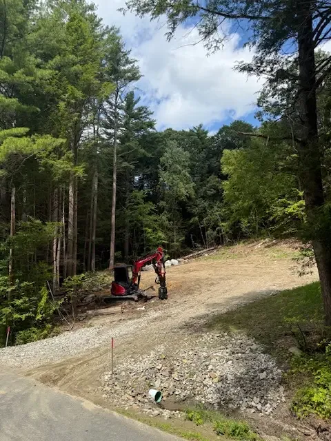 Red mini excavator clearing a path through a wooded lot beside a gravel pile