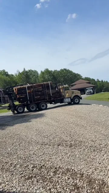 Log truck loaded with timber turning onto a gravel driveway beside a country home