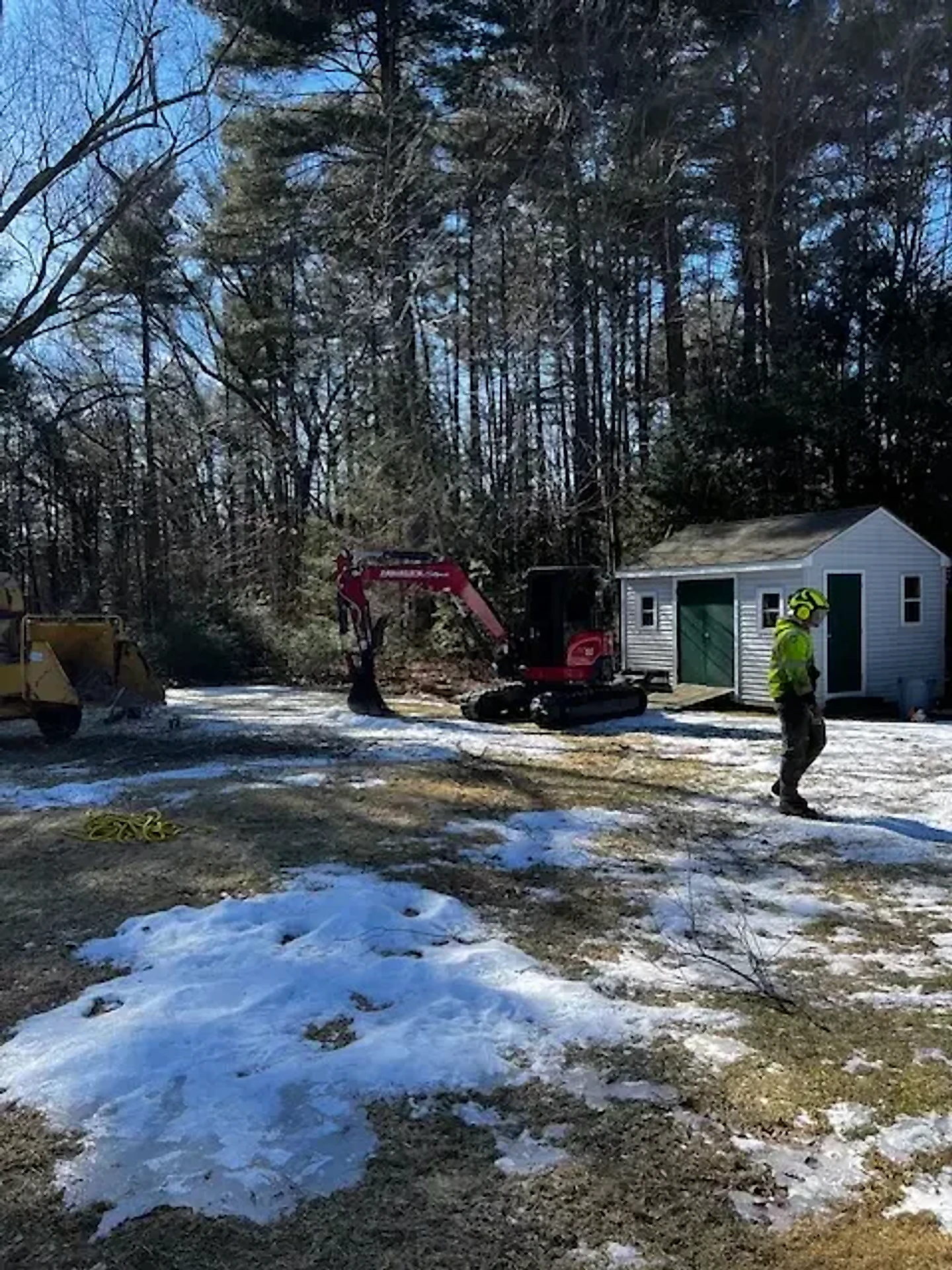 Red mini excavator and a crew member in a hi-vis jacket working beside a home on a snowy lot, ready for septic and trenching work