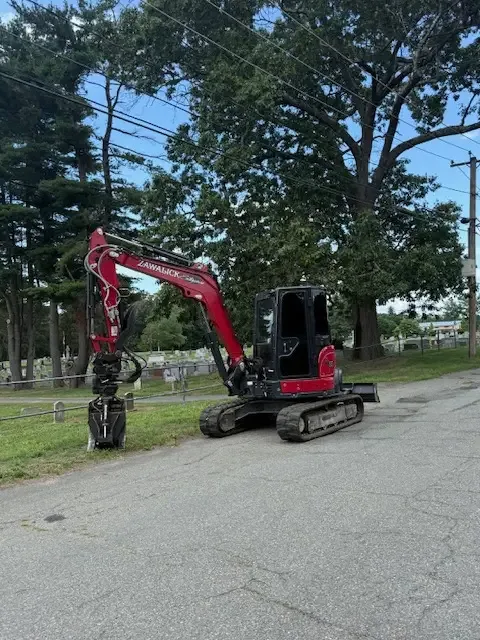 Red mini excavator with a grapple attachment parked beside a road under tall pines