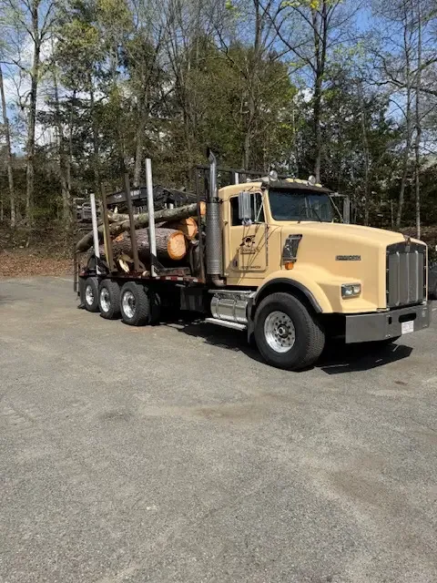 Tan log truck with a stack of cut logs on the bed, parked behind a bucket truck