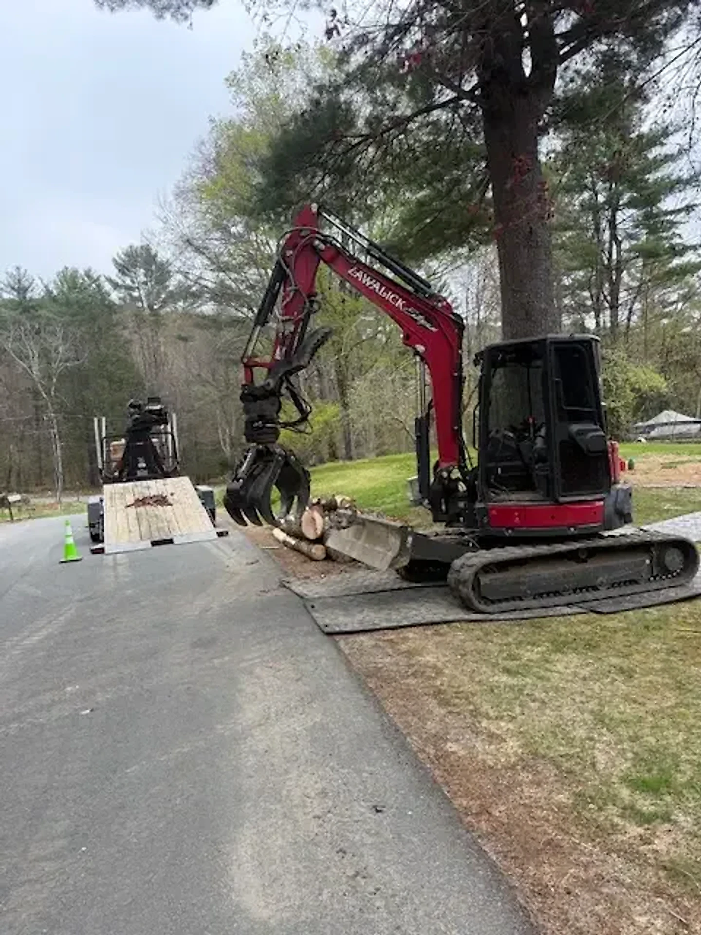 Red Zawalick excavator with a grapple attachment on a paved driveway beside a lawn and tall pines